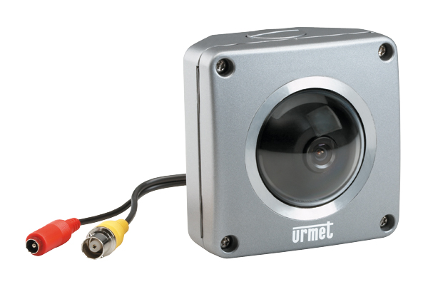 Kompakte Farb-Mini-Kamera von Urmet in einem quadratischen, silbernen Gehäuse mit schwarzer Kuppeloptik. Zwei Kabelanschlüsse mit gelbem und rotem RCA-Stecker für Video- und Stromverbindung.