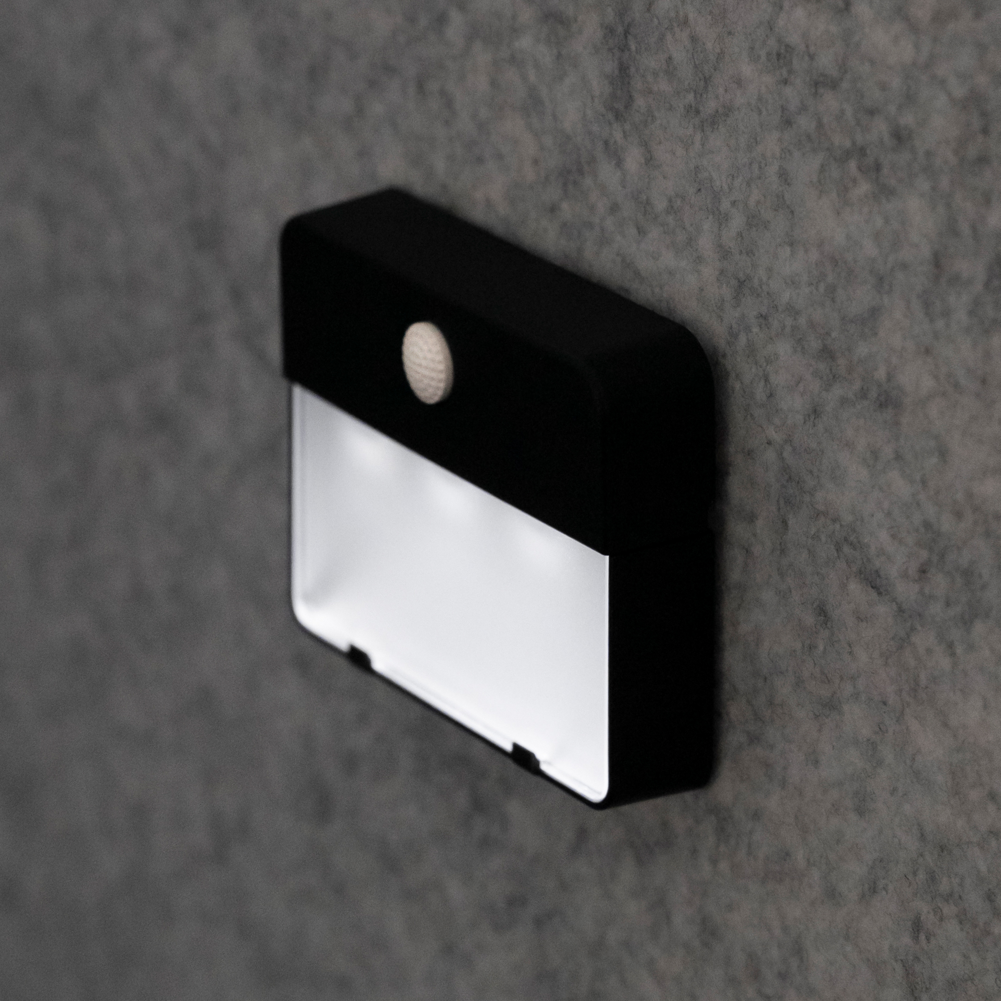 Klingelplatte in Schwarz mit beleuchtetem Namensfeld, Seitenansicht an grauer Wand montiert, LED-Licht gut sichtbar.